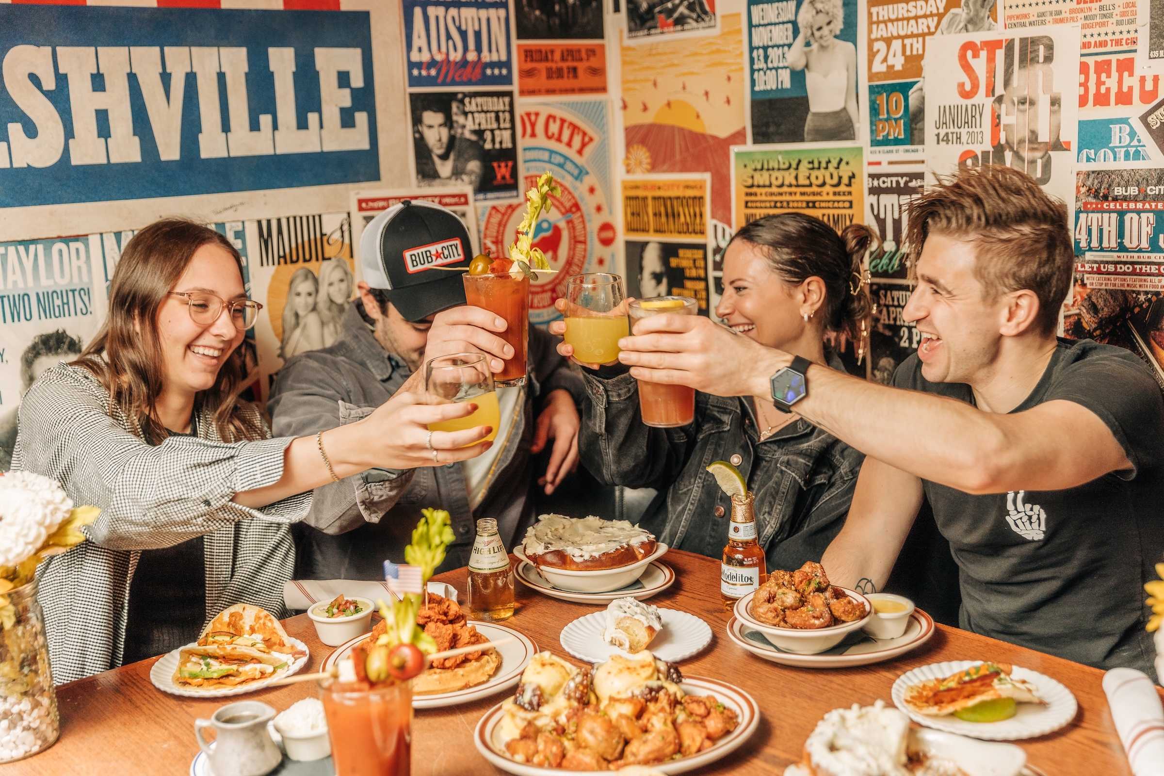 4 people sit at a table in a bright area with southern-style music posters on the wall behind them. The table is filled with brunch food and the people are raising their glass in a celebratory cheers.